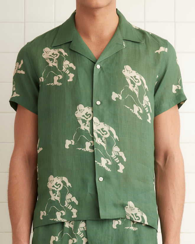 Green linen short sleeve shirt with images of football players across the garment. Four button down front seam with pointed collar.