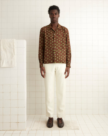 In a white-tiled room, a person gazes directly at the camera, wearing the Hop Tree Long Sleeve Shirt with a 1930s-inspired brown patterned block print crafted from cotton voile, paired with white pants.