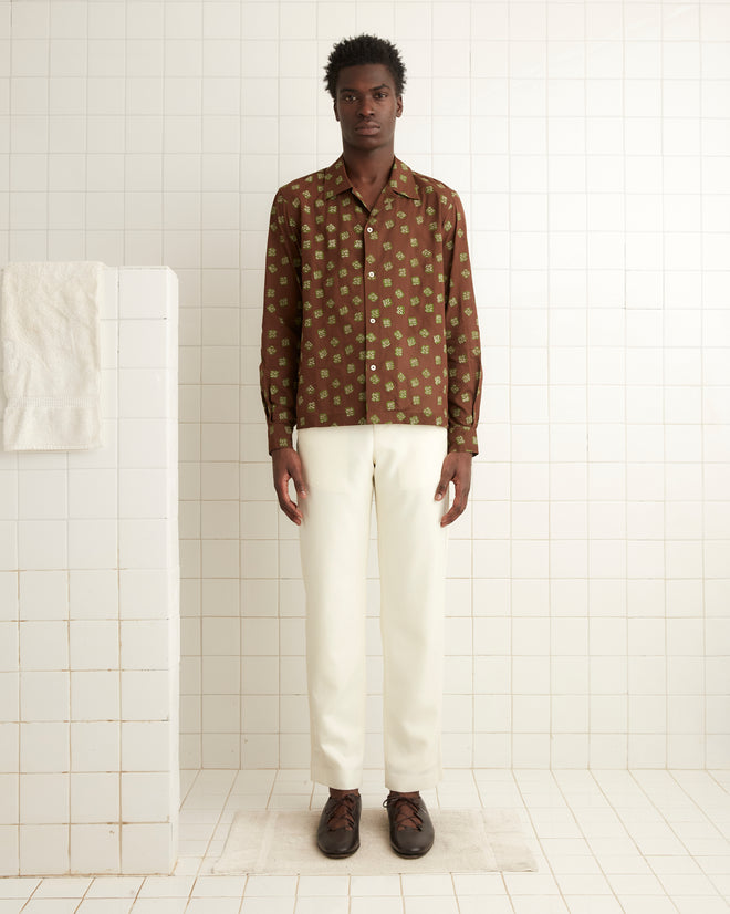 In a white-tiled room, a person gazes directly at the camera, wearing the Hop Tree Long Sleeve Shirt with a 1930s-inspired brown patterned block print crafted from cotton voile, paired with white pants.