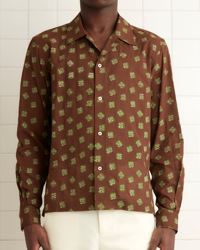 In a white-tiled room, a person gazes directly at the camera, wearing the Hop Tree Long Sleeve Shirt with a 1930s-inspired brown patterned block print crafted from cotton voile, paired with white pants.