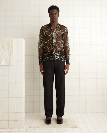 A person wearing a Sheer Fête Shirt embellished with sequins and floral designs, paired with black pants, stands on a white tiled floor.