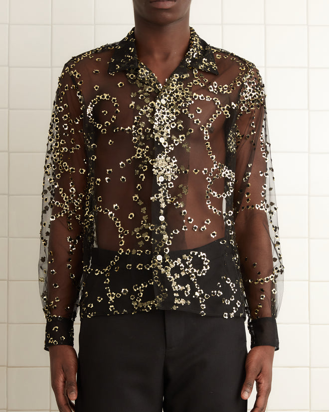 A person in a Sheer Fête Shirt adorned with delicate sequins and paired with black pants stands in a tiled room with a towel hanging nearby.