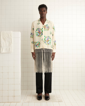 A person dressed in a Flower Language Shirt and a fringed skirt stands in a tiled room, with a towel on the left.