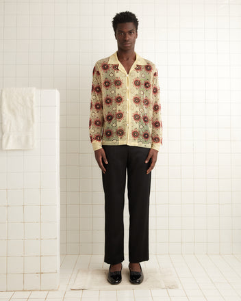 A person wearing a Solarium Long Sleeve Shirt and black pants stands in a white tiled room, exuding the elegance of the 1930s. Beside them, a towel adorned with intricate handmade beadwork hangs on a wall hook, subtly reflecting the charm of that era.