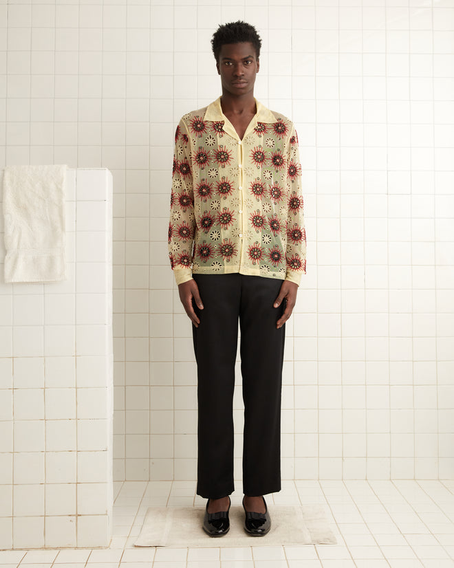 A person wearing the Solarium Long Sleeve Shirt featuring handmade beadwork and black pants stands in a tiled room next to a towel on a rack.