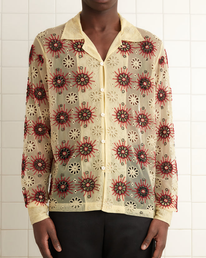 A person wearing the Solarium Long Sleeve Shirt featuring handmade beadwork and black pants stands in a tiled room next to a towel on a rack.