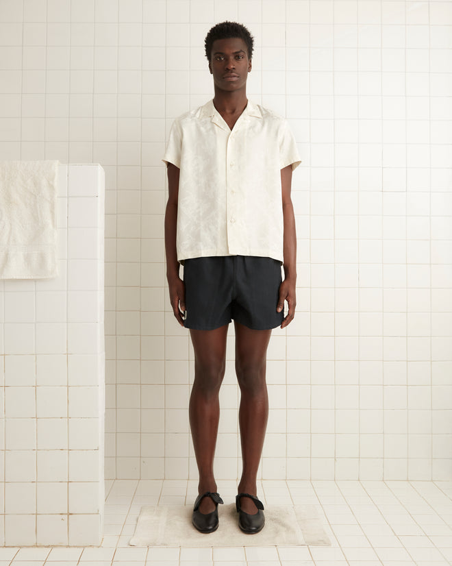 Person stands in a tile-walled room wearing a Football Jacquard Bowling Shirt - Cream, black shorts, and black shoes.