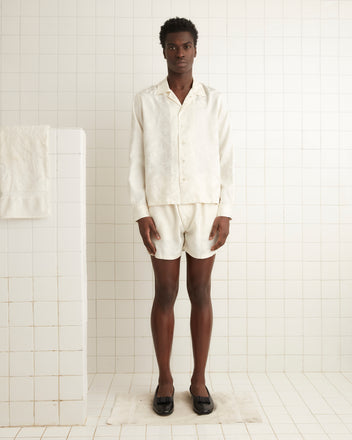 A person stands in a white-tiled room wearing the Football Jacquard Havana Shirt - Cream, paired with black slippers.