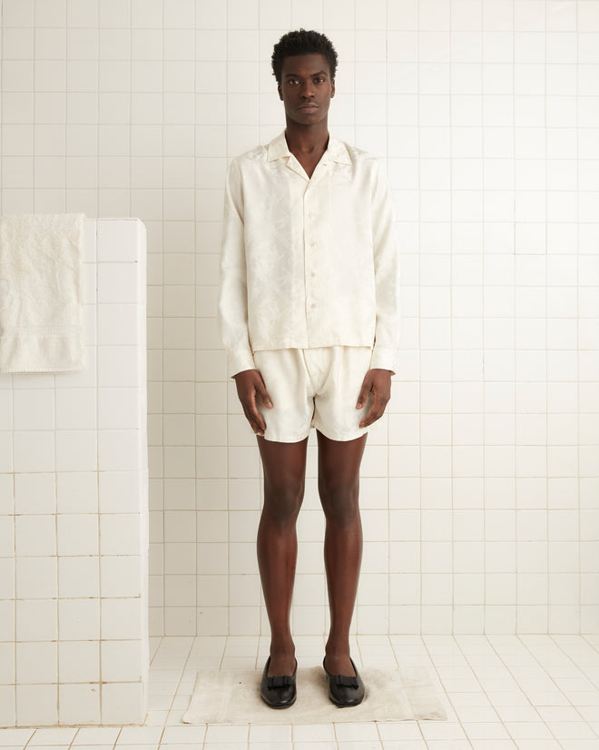 A person stands in a white-tiled room wearing the Football Jacquard Havana Shirt - Cream, paired with black slippers.