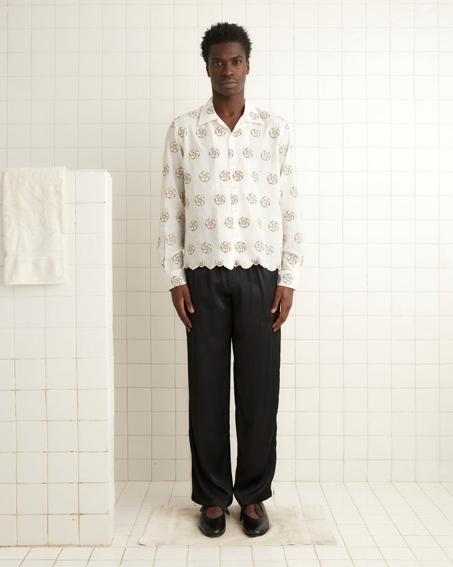 A person stands in a tiled room, wearing the Floral Aperture Long Sleeve Shirt paired with black pants. A towel hangs on the tiled wall, enhancing the room's simple elegance.