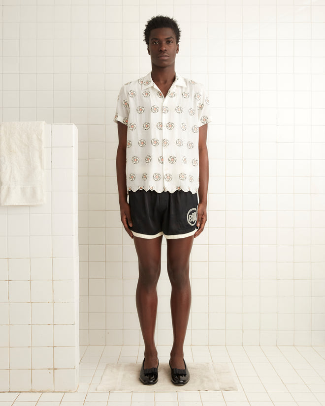 A person stands in a tiled room, wearing the Floral Aperture Short Sleeve Shirt featuring geometric flower embroidery, paired with black shorts and black shoes. To the left, on the white wall, hangs a towel.