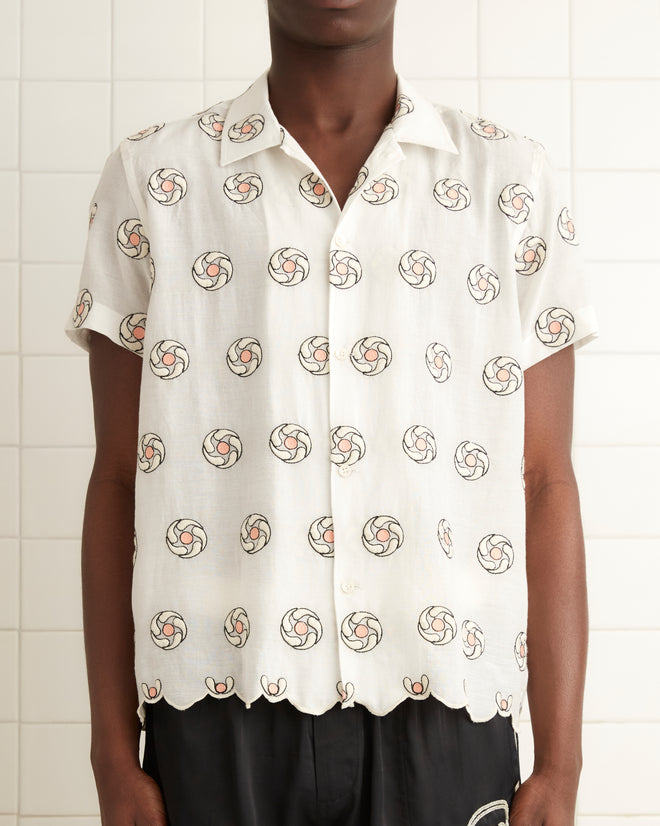 A person stands in a tiled room, wearing the Floral Aperture Short Sleeve Shirt featuring geometric flower embroidery, paired with black shorts and black shoes. To the left, on the white wall, hangs a towel.