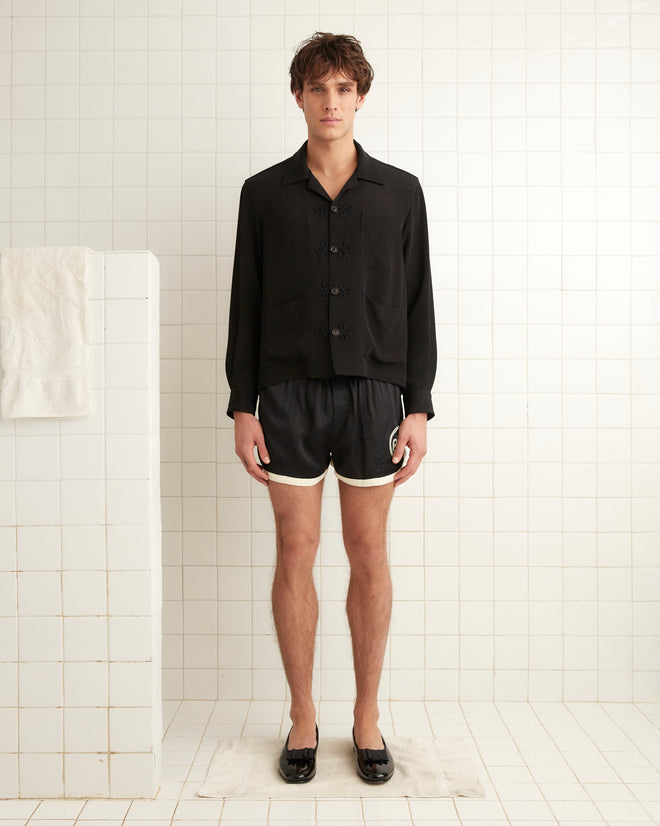 In a tiled room, an individual is seen wearing the Trillium Long Sleeve Shirt in black, evoking 1940s men's fashion. They are also dressed in black shorts with white trim and black shoes. A white towel is hanging on the tiled wall next to them.