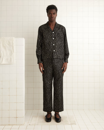 A man wearing a black Glitter Shirt stands in a white tiled bathroom with a towel hanging nearby.