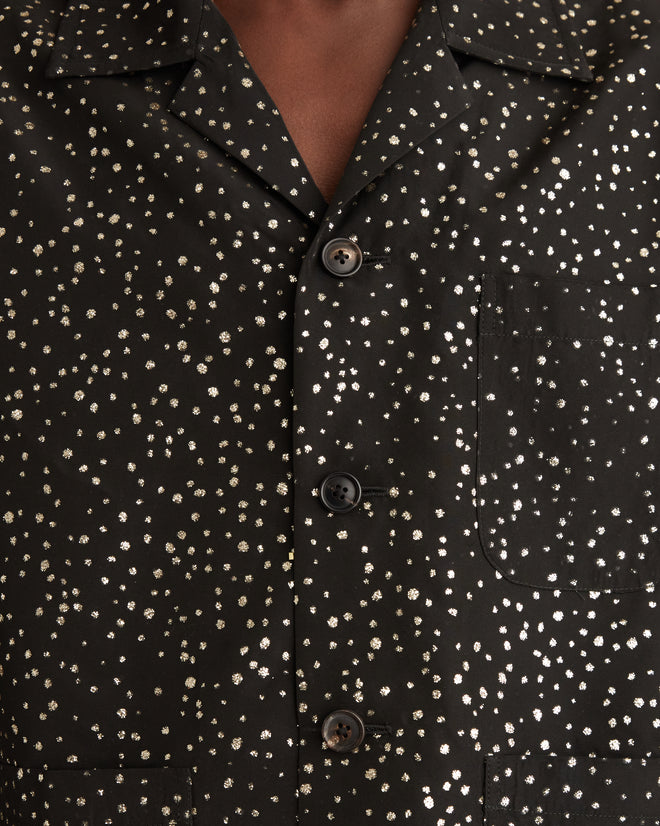 A person wearing a black Glitter Shirt stands in a white tiled room with a towel on the wall.