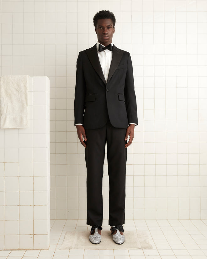 A person in a Barathea Peak Lapel Tuxedo Jacket sits in front of a piano, with a vase of flowers gracing the background.