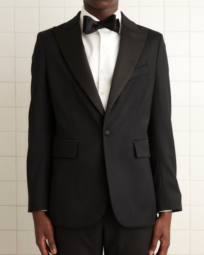 A person in a Barathea Peak Lapel Tuxedo Jacket sits in front of a piano, with a vase of flowers gracing the background.