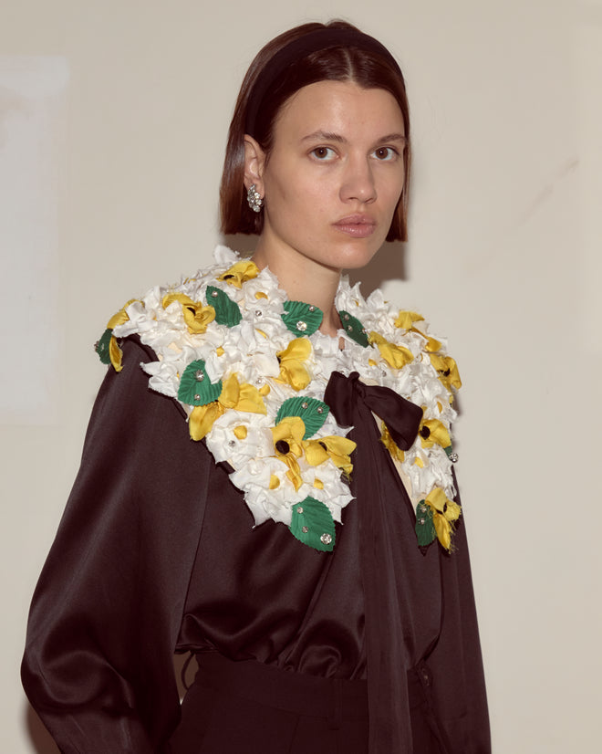 A woman indoors wears the Blossom Collar—a large floral accessory featuring white, yellow, and green flowers with a ribbon tie—styled over a black outfit.