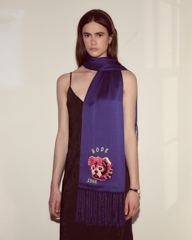 A woman in a black dress wears the Dream Souvenir Scarf, a textured silk accessory embroidered with "BODE 1966" and a dog motif.