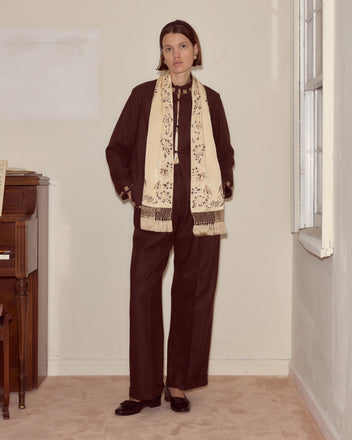 Dressed in a dark suit, a person stands indoors beside a piano and sunlit window, wearing the Phantasm Fringe Scarf with intricate Victorian-inspired embroidery.