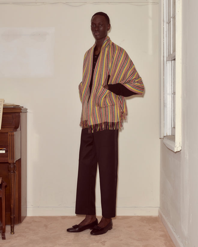 A person stands indoors by a piano and window, dressed in black pants, a black top, and the Lawrence Shawl—a vibrant merino piece with bold stripes that adds an eye-catching finish to their outfit.