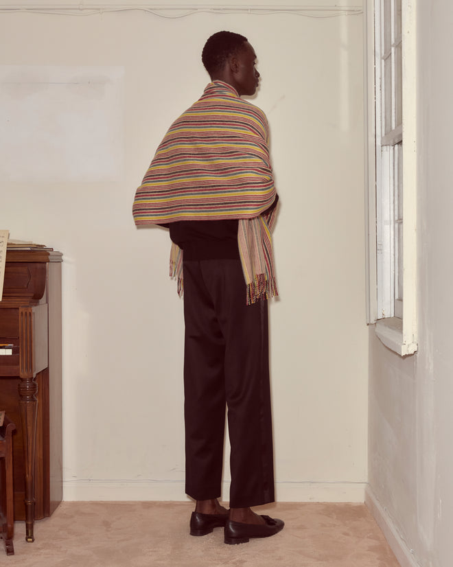 A person in black pants and a Lawrence Shawl featuring bold stripes stands facing a corner near a piano and window.