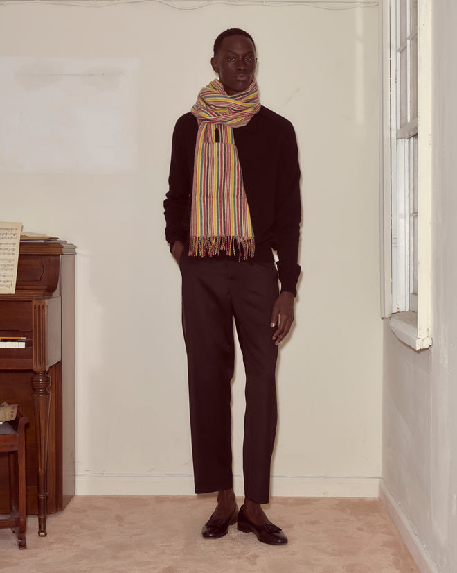 A man in black clothing stands indoors by a piano and window, showcasing his unique Lawrence Shawl—a colorful merino accessory with striking primary stripes.