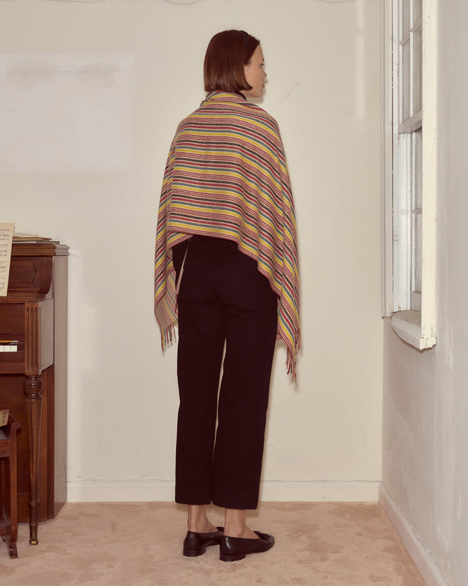 A person with short hair stands indoors, facing a wall, wearing the Lawrence Shawl with primary stripes and black pants.