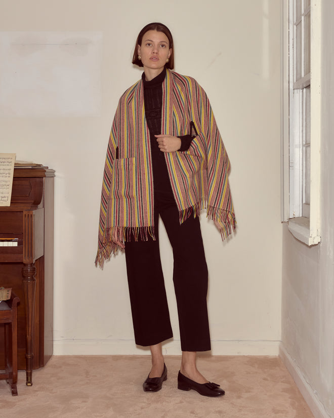 A woman wearing the Lawrence Shawl, featuring primary stripes, pairs it with a black outfit and stands indoors by a piano and window, looking at the camera.