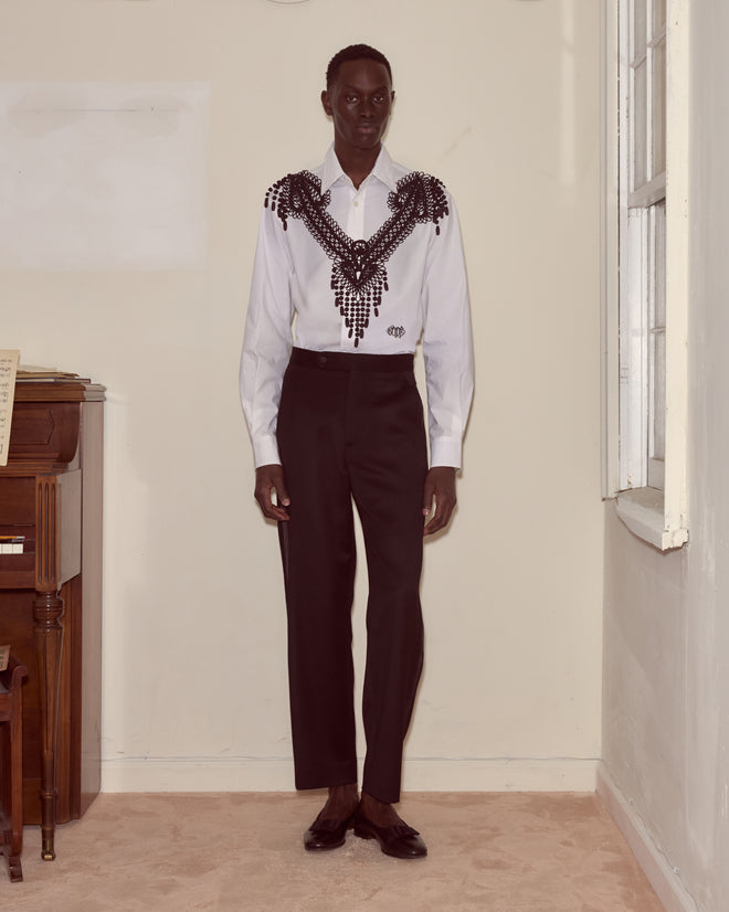 Indoors, a person wears a white shirt adorned with the Jet Braid Collar, featuring black beaded designs inspired by mourning jewelry, paired with black trousers.