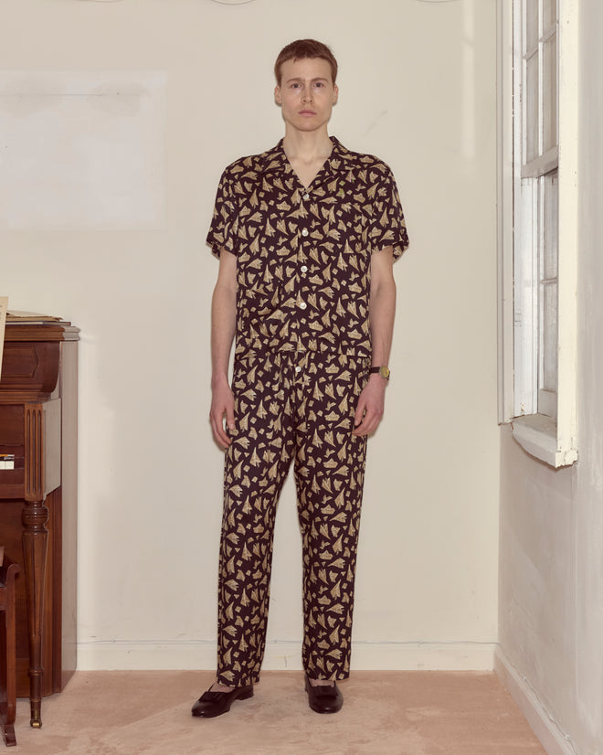 A man stands indoors wearing black Paper Hat Pajama Pants with a beige leaf novelty print and a matching shirt.