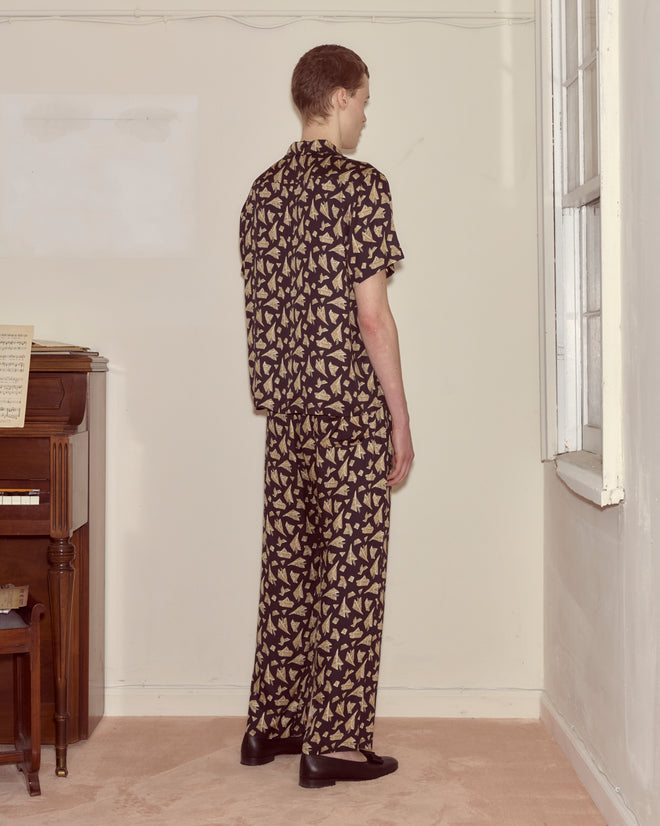 Wearing Paper Hat Pajama Pants with a matching print, a person stands by a piano in a softly lit room, facing away.