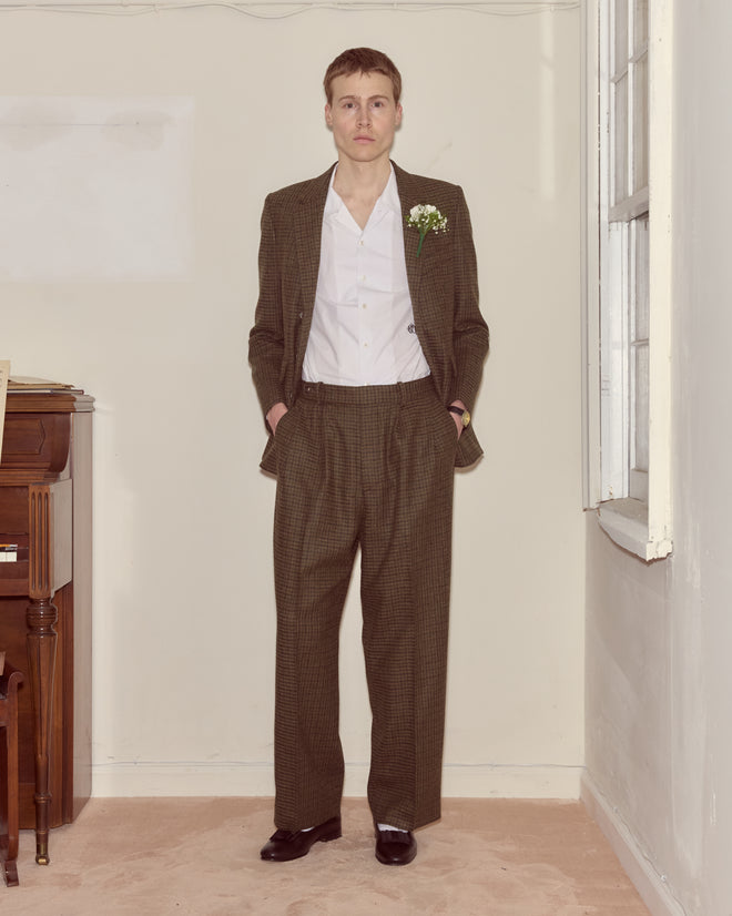 A person wearing Morris Houndstooth Trousers, a brown checked suit, white shirt, and boutonniere stands indoors near a window and piano.