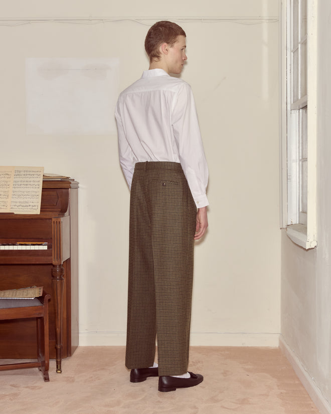 A person wearing a white shirt and Morris Houndstooth Trousers stands by a window, facing away, near a piano with sheet music.