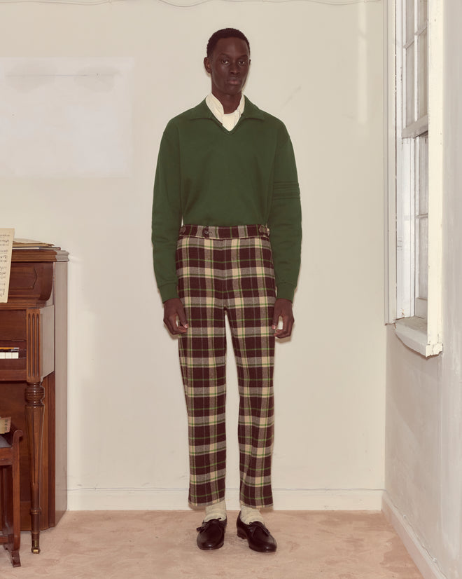 Indoors by a piano and window, a person wears a green sweater and Woodcrest Trousers—classic 100% wool plaid pants.
