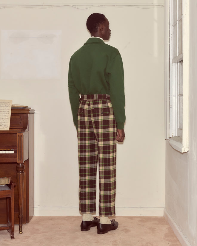 A person stands indoors, facing away, near a piano and window, wearing plaid Woodcrest Trousers and a green top.