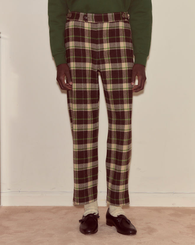 A person wearing Woodcrest Trousers in green and brown plaid 100% wool, paired with cream socks and black loafers, stands against a plain wall.