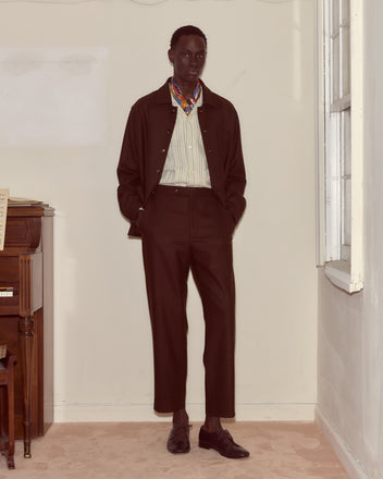 A man stands indoors next to a piano and window, wearing Bramble Side-Buckle Trousers paired with a dark suit jacket, striped shirt, and scarf.