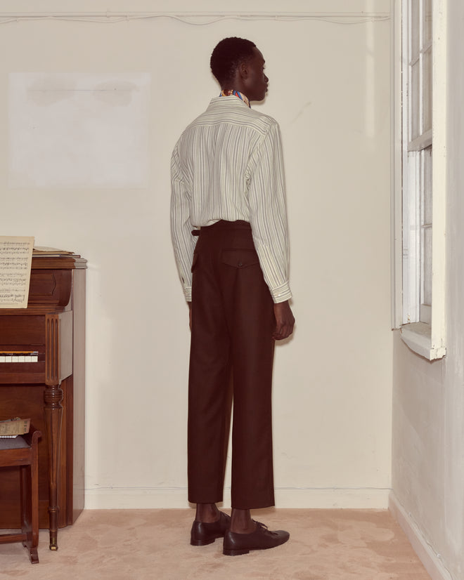 A person stands facing a beige wall in a room with a piano and window, wearing a striped shirt and Bramble Side-Buckle Trousers.