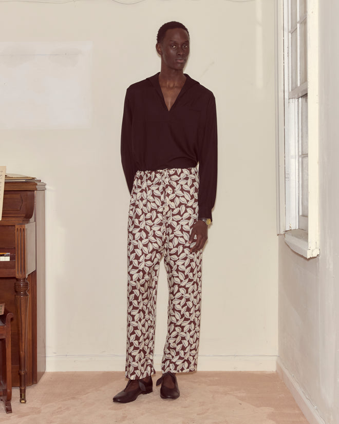 A man stands indoors wearing a black shirt and Paper Birch Pajama Pants, featuring a bold white leaf-stamp design with striking red patterns.