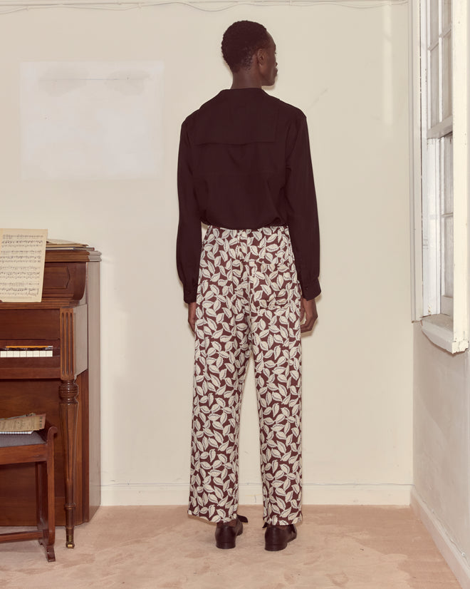 A person in a black shirt and Paper Birch Pajama Pants stands facing a wall beside a piano in a softly lit room.