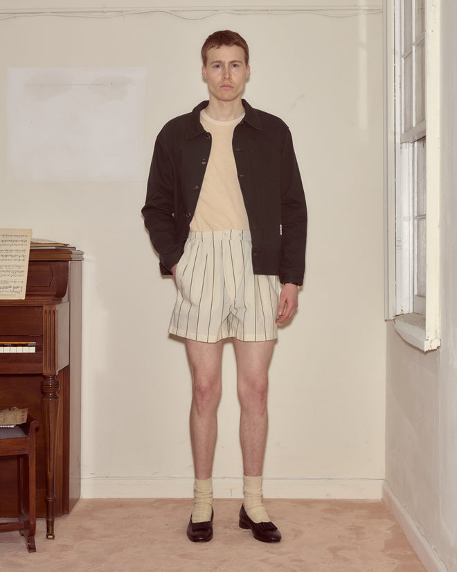 A man stands indoors wearing a black jacket, beige shirt, Celery Stripe Boxers made from 100% cotton, socks, and black shoes.
