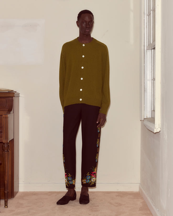 Indoors, a person wears an olive cardigan with Beaded Aquitaine Trousers—black felted wool featuring mid-rise straight legs and colorful hand-beaded embroidery at the hems.