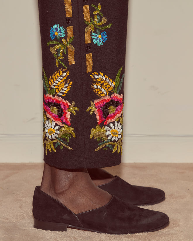 A person wearing black loafers and Beaded Aquitaine Trousers featuring heavyweight wool and colorful folk floral embroidery near the hems.