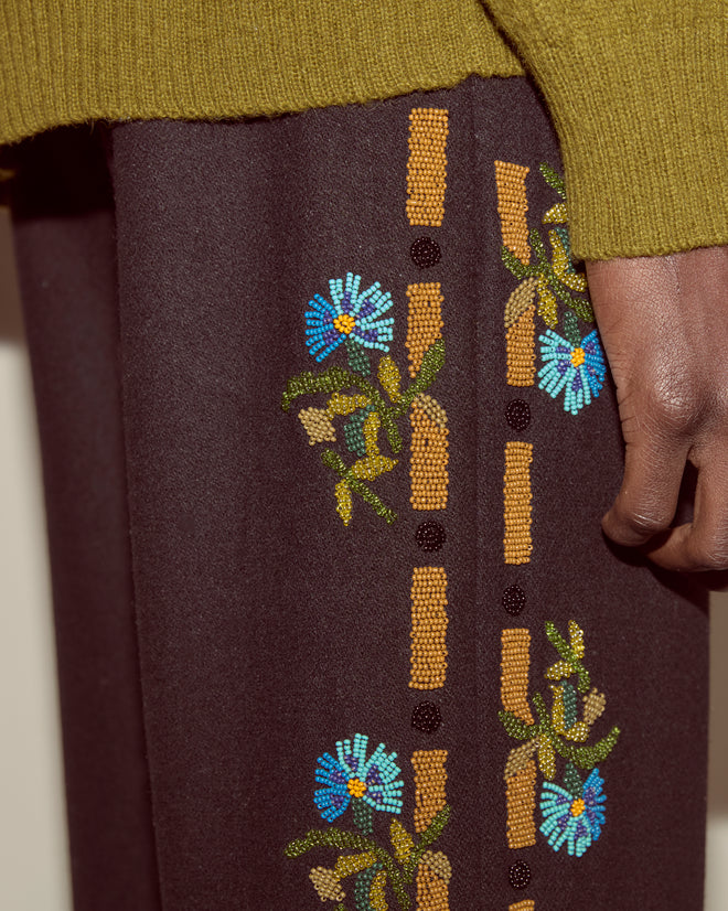 Close-up of a person wearing Beaded Aquitaine Trousers, featuring heavyweight wool with blue folk floral embroidery, paired with a green knit sweater.