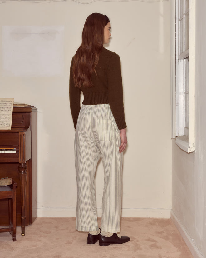 A woman with long hair stands indoors near a piano, facing a wall and wearing Electric Light Pajama Pants paired with a dark sweater.