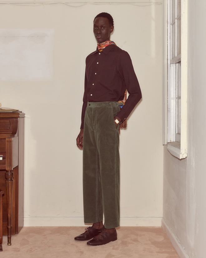 A person in a dark shirt, Corduroy Side-Buckle Trousers - Green, and a scarf stands indoors near a window and wooden furniture.