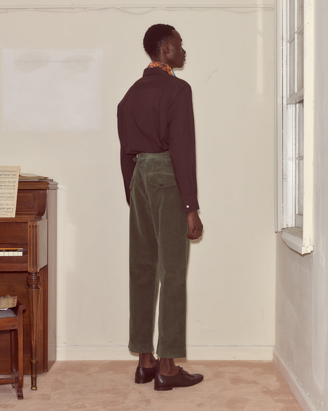 A person in a black shirt and green Corduroy Side-Buckle Trousers stands facing a wall near a piano and a window.