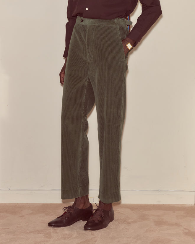 Person wearing Corduroy Side-Buckle Trousers - Green, a black shirt, and black shoes stands on a beige carpet.