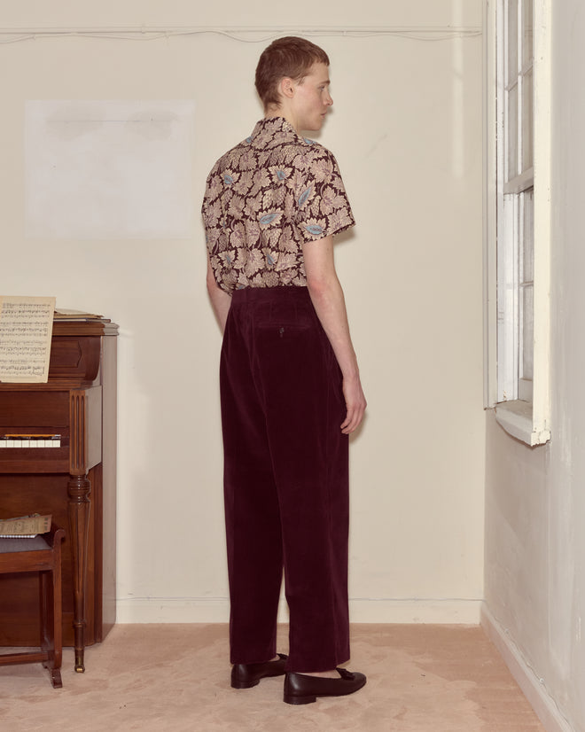 A person stands sideways in a plain room near a window and piano, wearing a patterned shirt and Corduroy McNab Trousers in dark purple with a wide-leg silhouette.